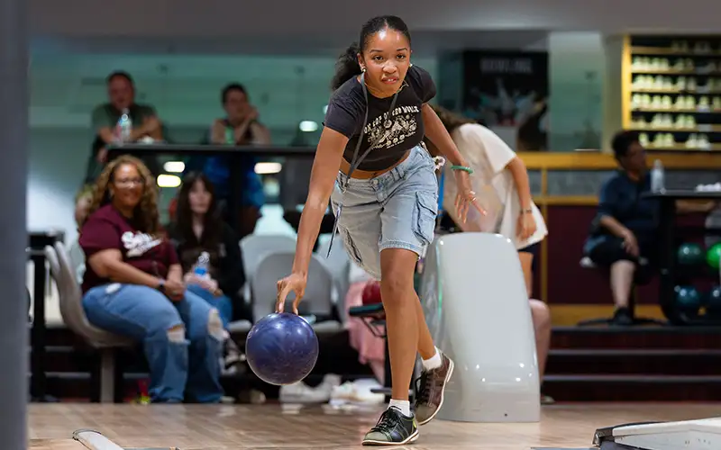 Student bowling at a campus alley, mid-action with a focused expression, while others watch and socialize in the background.
