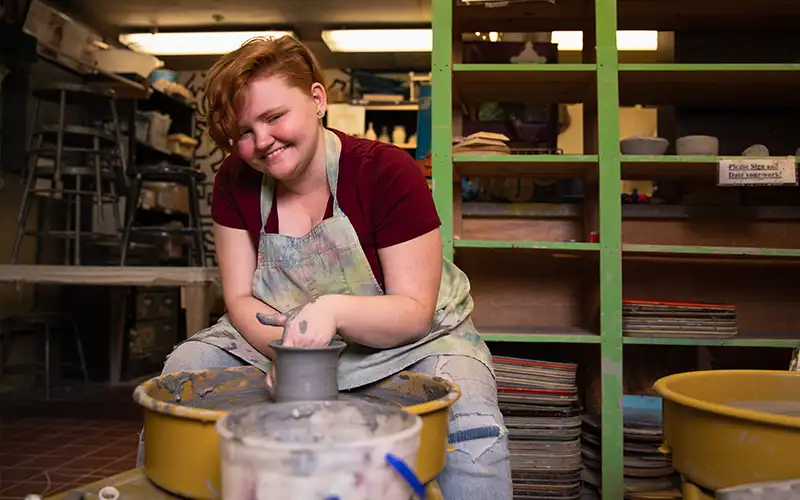 Student smiling while shaping clay on a pottery wheel in a campus craft studio, wearing an apron and surrounded by shelves with ceramic pieces and supplies.