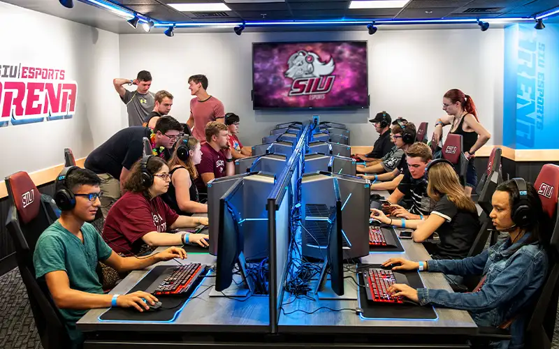 Students competing in an esports match at the SIU Esports Arena, focused on gaming monitors with illuminated keyboards and a large SIU logo screen in the background.