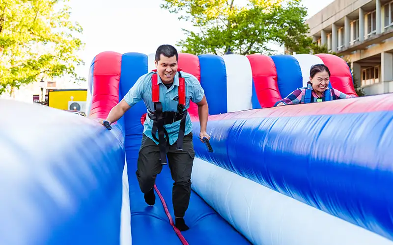 Two students race side by side on an inflatable bungee run during a campus event, smiling and wearing harnesses as they compete.