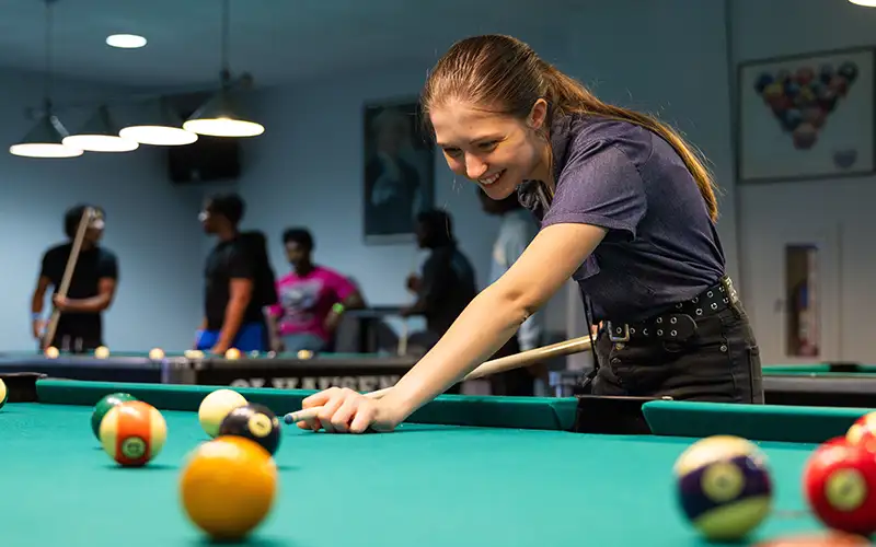 Student smiling while aiming a shot during a game of billiards in a campus recreation room, with other students socializing in the background.