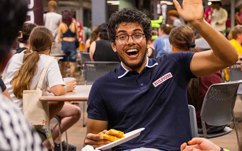 Student smiling and waving while holding a plate of pizza in a busy campus dining area filled with students socializing and eating.