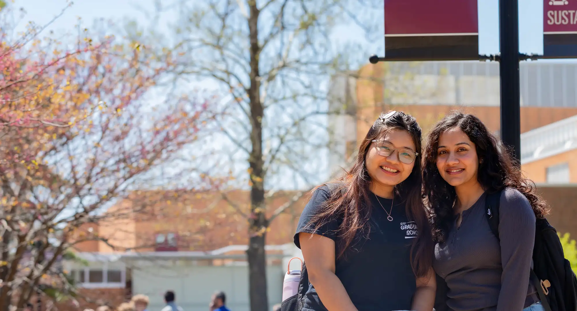 Two students smile outdoors on campus during spring, wearing backpacks and casual clothes, with budding trees and other students in the background.