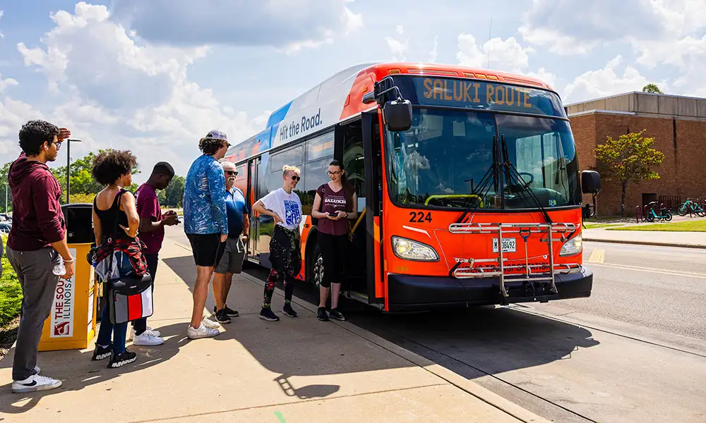 Students wait to board a bright orange Saluki Route bus on campus under a partly cloudy sky, with bicycles and a brick building in the background.