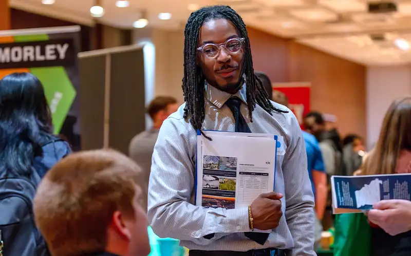 Student dressed in business attire holding a resume folder at a career fair, engaging in conversation with a recruiter in a busy event space.