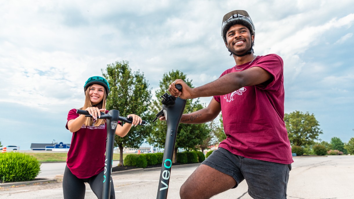 two students smiling as they stand on the scooter